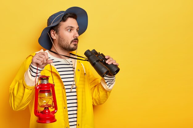 Serious unshaven man observes something with binoculars, holds red oil lamp, walks on foot, explores new place, wears hat and raincoat, isolated over yellow wall