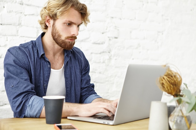 Serious unshaven young European journalist sitting at wooden cafe table keyboarding on modern laptop, looking for important information on Internet while working on article for online paper