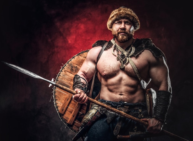 Serious Viking clad in light armor with a shield behind his back holds a spear. Posing on a dark background with red light