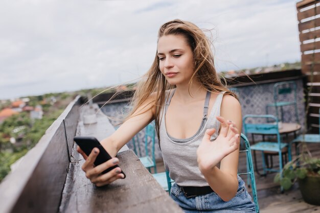 Серьезная белая девушка, делающая селфи в ветреный весенний день. Открытый выстрел довольной европейской молодой женщины позирует с телефоном в кафе на крыше.