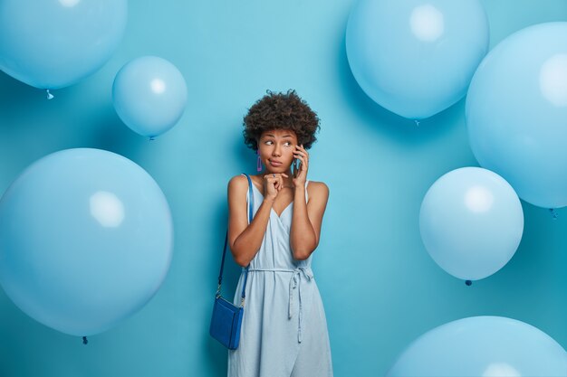 Serious woman calls friend, holds smartphone near ear, going to have walk in park, wears blue dress and bag to fit outfit, invites someone on party, prepares for celebration, stands near balloons