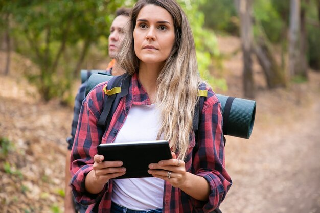 Serious woman checking path via tablet and walking on mountainous trail. Caucasian hikers or travelers carrying backpacks and hiking in forest. Backpacking tourism, adventure and vacation concept