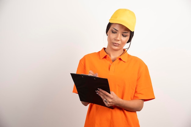 Serious woman courier with pencil looking on clipboard on white wall.