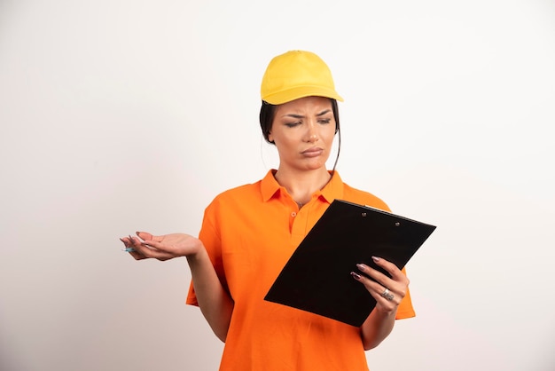 Serious woman courier with pencil looking on clipboard on white wall