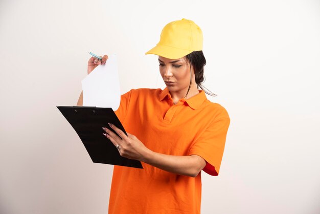 Serious woman courier with pencil looking on clipboard on white wall.