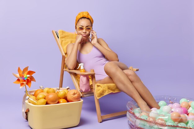 Serious woman dressed in swimsuit has phone conversation keeps cellular near ear poses on deck chair enjoys summer time sits on deck chair isolated over purple background Perfect vacation and rest