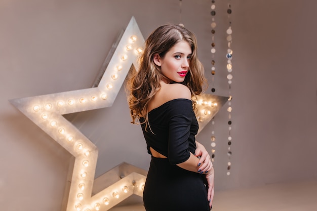 Serious woman in elegant outfit looking away standing on dark wall. Indoor portrait from back of shapely curly girl waiting for holiday event.