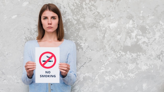 Serious woman holding with no smoking text and sign standing against weathered wall