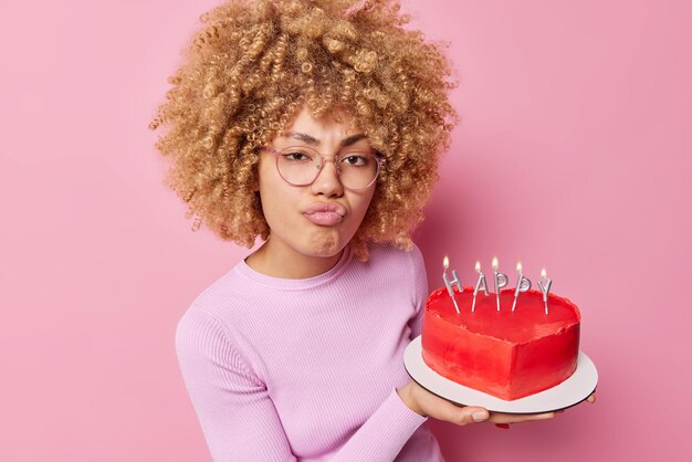 Serious woman makes unhappy grimace purses lips holds tasty heart shaped cake prepares for Valentines Day or birthday celebration wears transparent glasses casual jumper isolated over pink wall