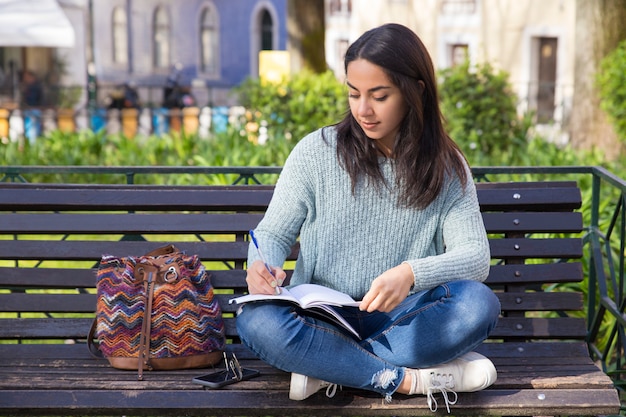 Serious woman making notes and sitting on bench outdoors