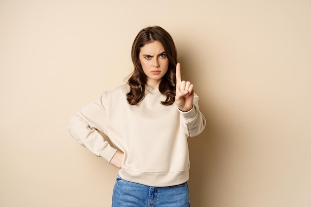 Free photo serious woman show one finger stop disapproval sign forbid smth say no prohibit standing over beige ...