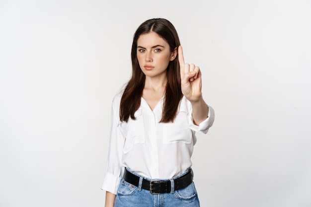 Серьезная женщина показывает табу, стоп-жест, неодобрительно трясет пальцем, не соглашается, что-то запрещает, стоит на белом фоне.