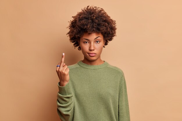 Serious woman shows fuck you sign looks with poker face being vulgar and has quarrel with someone dressed in casual sweater isolated over beige wall