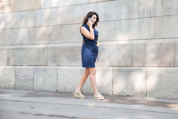 Serious woman walking along wall with cellphone