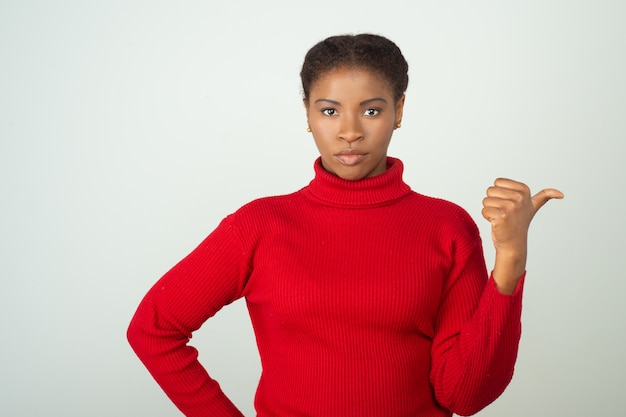 Serious woman wearing red sweater and pointing thumb to the side