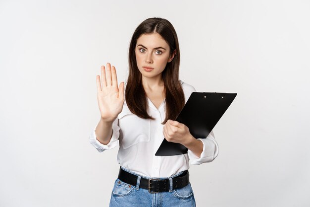 Serious woman with clipboard documents, extending arm, showing taboo stop sign, disapprove, prohibit smth, standing over white background