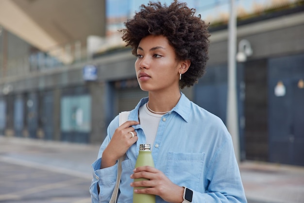 Serious woman with curly bushy hair hold bottle of fresh water wears denim shirt looks away pensively walks outdoors against blurred background waits for someone at street thinks about life