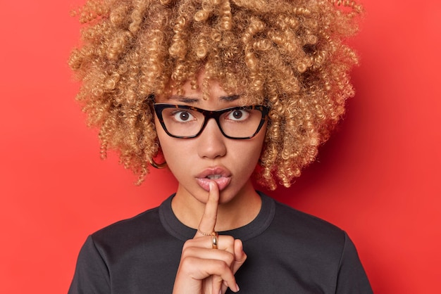 Serious woman with curly bushy hair keeps index finger over lips has mysterious expression makes hush gesture wears transparent glasses casual black t shirt isolated over red background Body language