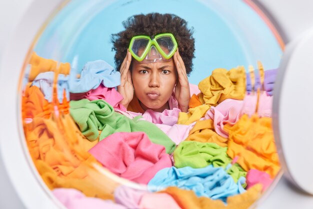 이마에 스노클링 마스크를 쓴 진지한 여성이 세탁기 보기에서 펼쳐진 다색 세탁물을 통해 정면 포즈에서 화난 표정으로 입술을 접은 채로 유지합니다