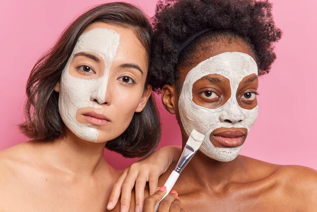 Serious women apply beauty masks on face do rejuvenation procedures at beauty salon pose with bare shoulders indoor look directly at camera stand against pink background. Skin care and wellness