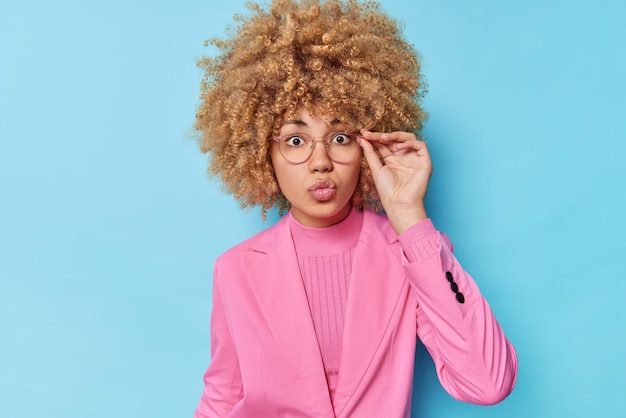 Serious wondered woman with curly bushy hair keeps lips folded hand on rim of spectacles dressed in formal outfit reacts on shocking news isolated over blue background Human face expressions