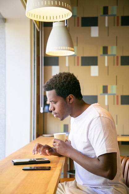 Serious young African American man drinking coffee while sitting at desk in co-working space, using tablet, typing and reading on screen