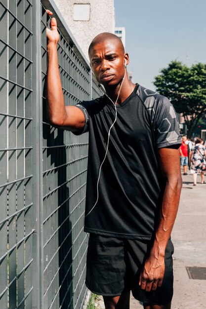 灰色の門の前に立っている彼の耳にイヤホンを持つ深刻な若いアフリカのオスの運動選手