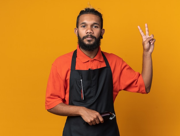 Serious young afro-american male barber wearing uniform holding hair clippers doing peace sign 