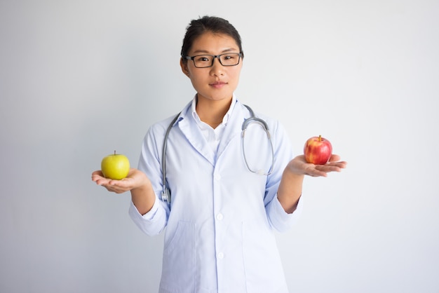 Serious young asian female doctor holding red and yellow apple.