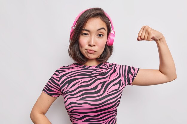 Serious young Asian woman has determined face expression raises arm shows biceps feels strong listens music via wireless headphones dressed in striped t shirt isolated over white wall