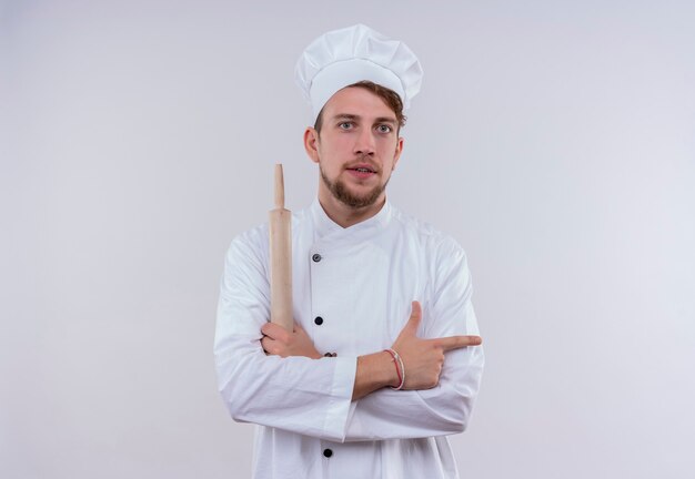 A serious young bearded chef man wearing white cooker uniform and hat holding rolling pin and pointing side while looking on a white wall