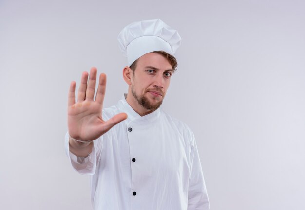 A serious young bearded chef man wearing white cooker uniform and hat showing stop gesture with hand while looking on a white wall