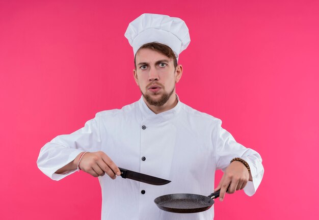 Серьезный молодой бородатый шеф-повар в белой форме держит сковороду с ножом, глядя на розовую стену