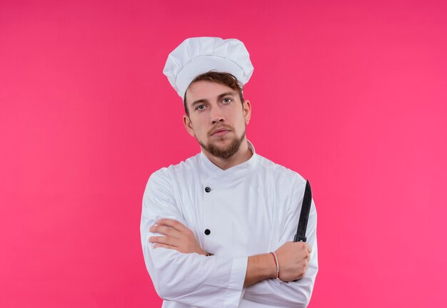 A serious young bearded chef man in white uniform holding a knife while looking on a pink wall