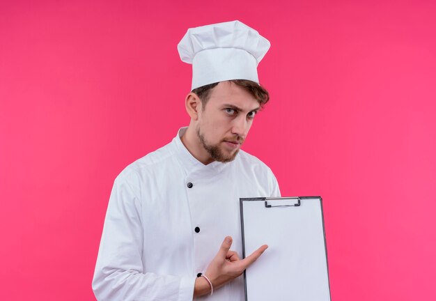 분홍색 벽을 보면서 빈 폴더를 가리키는 흰색 제복을 입은 심각한 젊은 수염 난 요리사 남자