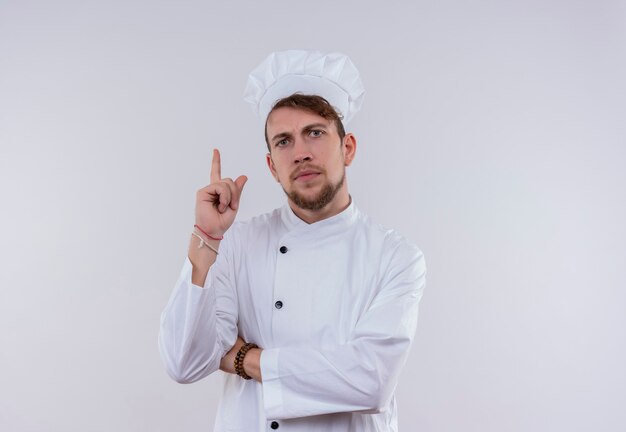 흰 벽에보고있는 동안 접힌 손으로 가리키는 흰색 유니폼에 심각한 젊은 수염 난 요리사 남자