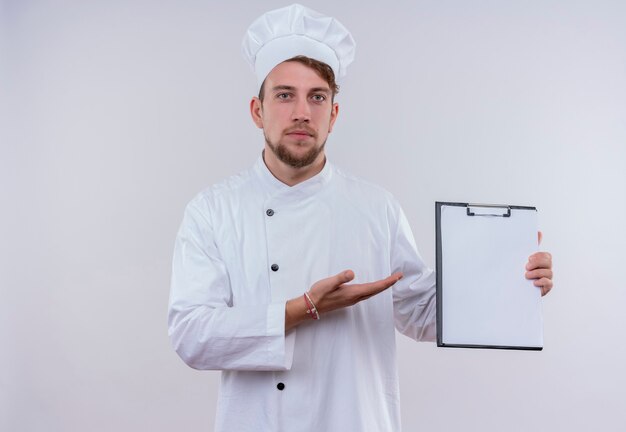 A serious young bearded chef man in white uniform showing blank folder while looking on a white wall