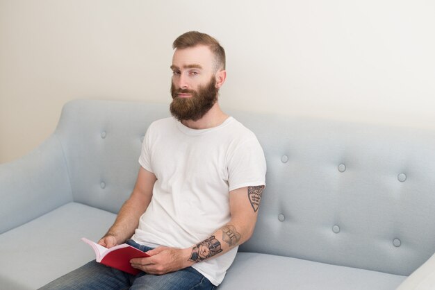 Серьезный молодой бородатый человек сидит на диване и читает книгу
