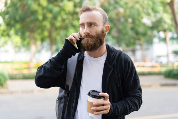 Serious young bearded man walking in city and calling on phone