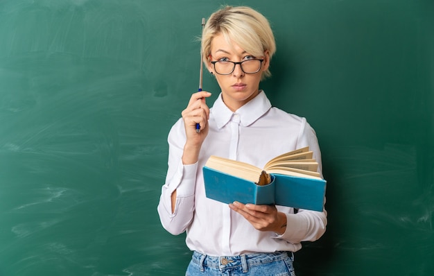 serious young blonde female teacher wearing glasses in classroom standing in front of chalkboard holding book touching head with pointer stick looking at front with copy space
