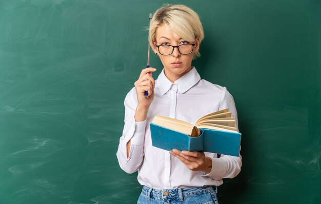 serious young blonde female teacher wearing glasses in classroom standing in front of chalkboard holding book touching head with pointer stick looking at front with copy space