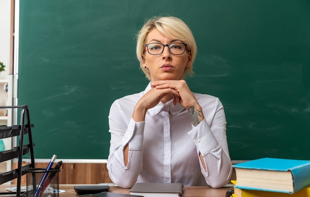Серьезная молодая блондинка учительница в очках сидит за столом со школьными инструментами в классе, держа руки вместе под подбородком, глядя в камеру