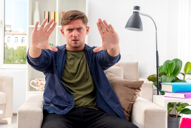 Serious young blonde handsome man sits on armchair gesturing stop hand sign with two hands inside the living room