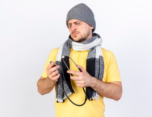 Serious young blonde ill man wearing winter hat and scarf holds and looks at stethoscope isolated on white wall