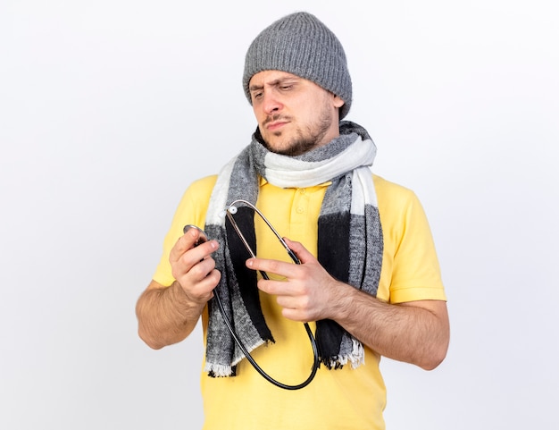 Serious young blonde ill man wearing winter hat and scarf holds and looks at stethoscope isolated on white wall