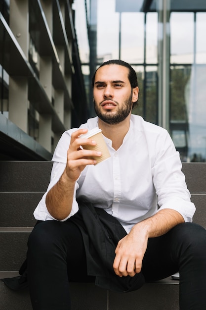 Serious young businessman sitting on staircase holding takeaway coffee cup