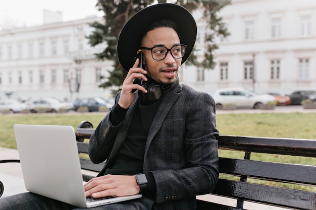 공원에서 컴퓨터와 함께 작업하는 동안 전화 통화하는 심각한 젊은 사업가. 광장에서 노트북을 사용하는 바쁜 아프리카 남자의 야외 사진.