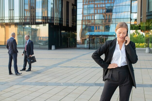 Serious young businesswoman in office suit talking on cellphone outdoors. Businesspeople and city building glass facade in background. Copy space. Business communication concept