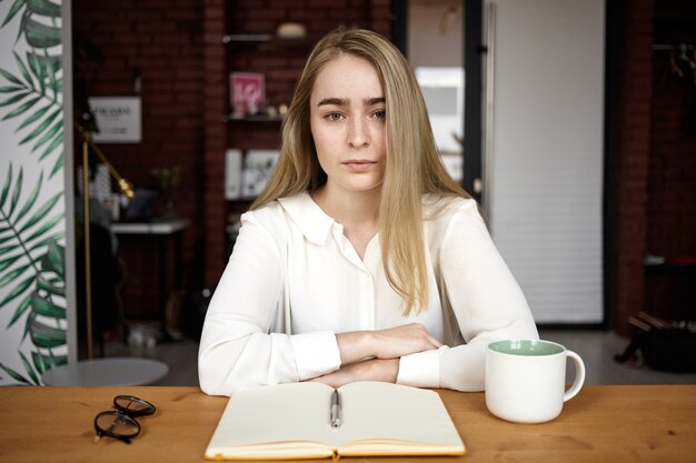 Serious young Caucasian female blogger sitting at desk with open notebook, glasses and mug, making notes while working on new article. People, lifestyle, job, occupation and creativity concept