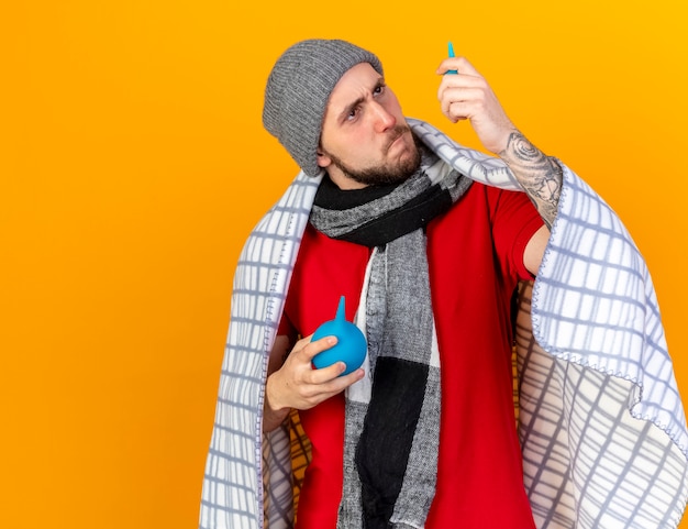 Serious young caucasian ill man wearing winter hat and scarf wrapped in plaid holds